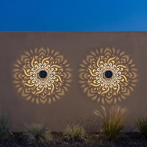 Peut inclure: Deux appliques solaires circulaires fixées sur une surface plane, projetant chacune un motif lumineux complexe et symétrique rappelant un mandala, diffusant une lueur dorée sur le mur sous un ciel bleu foncé.