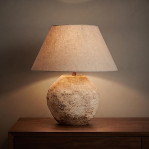 Op de afbeelding: Een tafellamp met een ronde, getextureerde, beige keramische voet met een gevlekte afwerking en een brede, conische, gebroken witte stoffen lampenkap. De lamp verspreidt een warme, zachte gloed onder de kap die de voet verlicht.