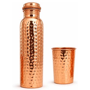 May include: A tall, cylindrical copper water bottle with a textured, hammered finish and a matching screw-top lid, accompanied by a smaller, matching hammered copper drinking cup. Both items feature a warm, metallic orange-brown colour.
