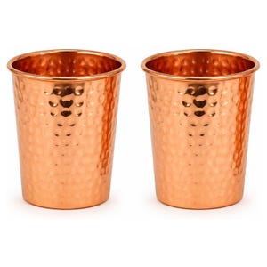 May include: A pair of identical cylindrical drinking cups crafted from polished copper. Each vessel features a distinct hammered texture across its exterior surface, creating a series of small, rounded indentations that catch the light.