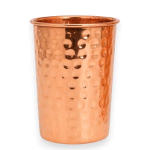 May include: A cylindrical copper tumbler featuring a distinct hammered texture across its entire exterior surface. The vessel has a smooth, rounded rim at the top and a polished metallic finish that reflects light, creating varied highlights.