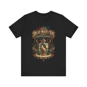 May include: A black short-sleeve t-shirt featuring a graphic print of a glass jar containing a toe, labeled Dead Man's Toe Apothecary & Oddities. The design includes ornate gold filigree, dark floral elements, moths, and the text Questionably Sourced, Wickedly Effective.