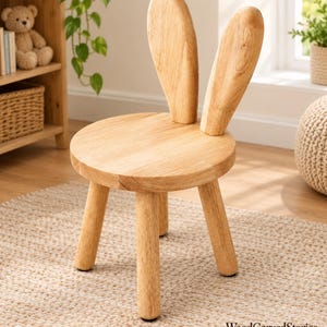 May include: A light-colored wooden chair featuring a round seat, four cylindrical legs, and a backrest shaped like two tall, upright rabbit ears. The natural wood grain is visible across the smooth surface of the chair, which stands on a textured rug.