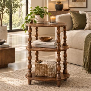 May include: A three-tiered round wooden side table with turned spindle legs. The top shelf holds a potted plant and a candle. The middle shelf features a wooden bowl and two books titled Kinfolk and At Home. A woven basket sits on the bottom shelf.