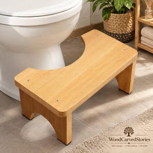 May include: A light brown wooden toilet stool with a curved cutout design, featuring four visible metal screw heads on the top surface and four sturdy legs with dark non-slip pads at the base, positioned in front of a white porcelain toilet.