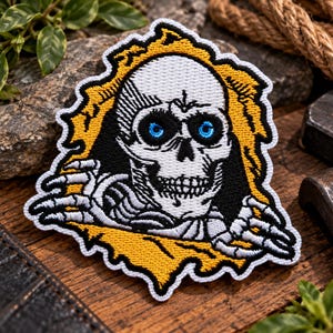 May include: An embroidered patch featuring a white skull with bright blue eyes and skeletal hands emerging from a jagged, golden-yellow border. The design includes black shading and a white outer edge, creating a high-contrast graphic.