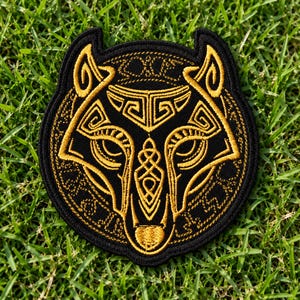 Op de afbeelding: Een ronde geborduurde patch met een gestileerd wolvenkopontwerp in gouddraad op een zwarte achtergrond. Het wolvengezicht is samengesteld uit ingewikkelde geometrische patronen en knoopwerk, met puntige oren en een centraal neusdetail.