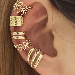 May include: A set of seven gold-toned ear cuffs worn along the cartilage and lobe. The collection includes two open-work leaf patterns, one textured band, two smooth double-wire bands, and two smooth single bands with a central beaded detail.