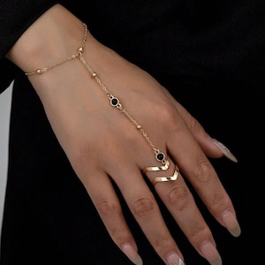 May include: A gold-toned slave bracelet featuring a delicate chain that connects a wrist band to a chevron-shaped ring. The chain is accented with small gold beads and two circular black stone settings positioned along the back of the hand.