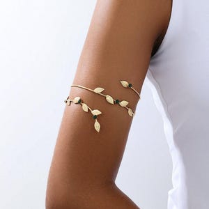 May include: A gold-toned arm cuff featuring a delicate vine design that wraps around the upper arm. The metal band is adorned with small, textured gold leaves and interspersed with tiny, round, dark green stones set along the vine structure.