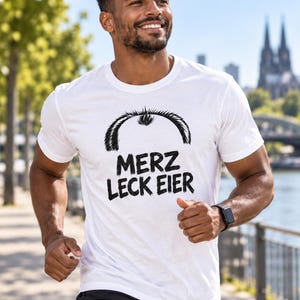 May include: A white short-sleeved t-shirt featuring a black graphic print of a receding hairline and the text "MERZ LECK EIER" in a bold, brush-stroke style font centered on the chest. The shirt has a classic crew neckline and a standard fit.