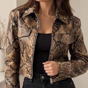 Peut inclure: Une veste courte avec un motif peau de serpent marron et noir, un col pointu, des manches longues et deux poches poitrine à rabat. La veste présente une fermeture éclair en métal argenté sur le devant et est portée sur un haut et un jean noirs.