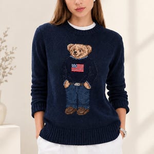 Può includere: Un maglione in maglia blu navy a maniche lunghe con una grafica centrale di un orsacchiotto che indossa un maglione con il disegno della bandiera americana, jeans blu e scarpe marroni. Il maglione presenta un girocollo, polsini e orlo a coste.