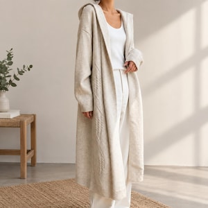 May include: A long, beige, open-front hooded cardigan featuring a subtle cable knit pattern along the front panels, worn over a white tank top and white trousers. The knit garment has long sleeves with ribbed cuffs and a relaxed, floor-length fit.