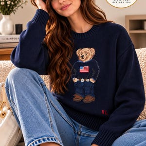 May include: A navy blue long-sleeved knit sweater featuring a central graphic of a teddy bear wearing a sweater with an American flag design, blue jeans, and brown shoes. The letters RL are embroidered in red on the lower left side of the sweater.