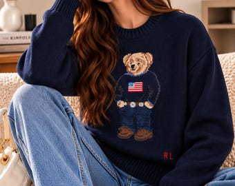 May include: A navy blue long-sleeved knit sweater featuring a central graphic of a teddy bear wearing a sweater with an American flag design, blue jeans, and brown shoes. The letters RL are embroidered in red on the lower left side of the sweater.