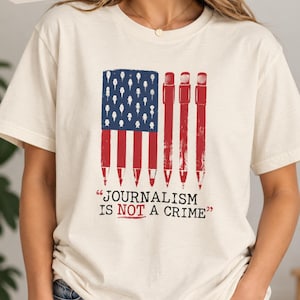 Puede incluir: Una camiseta de manga corta de color natural con un gráfico de una bandera estadounidense compuesta por lápices. Debajo del gráfico, el texto dice "JOURNALISM IS NOT A CRIME" con la palabra "NOT" subrayada en rojo. Diseño en rojo, blanco y azul.