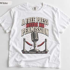 May include: A white short-sleeved t-shirt featuring a graphic of a vintage microphone between two stanchions with red ropes. The text reads A FREE PRESS NEEDS NO PERMISSION and FIRST AMENDMENT FREE PRESS in black and red lettering on the front.