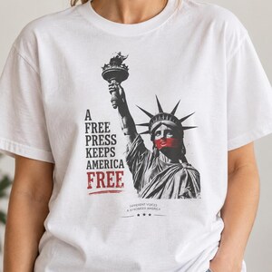May include: A white short-sleeve t-shirt featuring a black and white graphic of the Statue of Liberty with a red band over the mouth. The text reads A FREE PRESS KEEPS AMERICA FREE and DIFFERENT VOICES A STRONGER AMERICA with three stars.