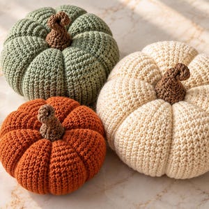 May include: Three crocheted pumpkins in sage green, burnt orange, and cream colors. Each pumpkin features a textured, ribbed surface and a small, brown, spiraled stem at the top. The items are arranged in a cluster, showcasing their round shapes.