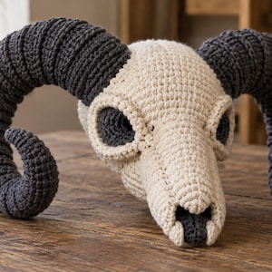 May include: A crochet ram skull sculpture featuring a cream-colored cranium and dark charcoal gray curved horns. The piece is crafted with visible yarn stitches, creating a textured surface that mimics the anatomical structure of a ram's head.