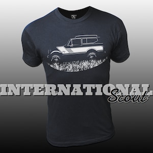 May include: A dark blue t-shirt featuring a white silhouette of an International Scout vehicle. The vehicle is depicted driving through grass. The text "INTERNATIONAL Scout" is printed below the vehicle.
