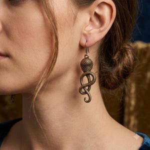 May include: A bronze-toned metal drop earring featuring a detailed, coiled snake design with a textured, scale-like surface. The snake is depicted in a vertical, winding position, suspended from a simple metal hook ear wire.