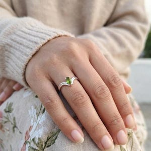 Könnte beinhalten: Ein Silberring mit einem marquise-geschliffenen grünen Edelstein in einer V-förmigen Ringschiene. Der Ring hat eine polierte metallische Oberfläche und eine Krappenfassung, die den zentralen Stein sichert und ein schlichtes Design bildet.