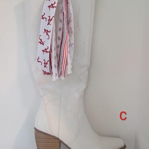 Könnte beinhalten: Ein weißer, kniehoher Westernstiefel mit Blockabsatz und dezenter Ton-in-Ton-Stickerei. An der Zugschlaufe hängt ein Bündel dekorativer Bänder mit roten Buchstaben-A-Aufdrucken, grauen Punkten sowie rot-weiß gestreiften Mustern. Der Buchstabe C ist zu sehen.