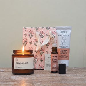 May include: A gift set featuring a lit amber glass candle labelled Morning Light, a pink floral patterned gift bag, a tube of Aery Hand Therapy Happy Space hand cream, and a small bottle of Aery Happy Space pulse point oil with a black cap.