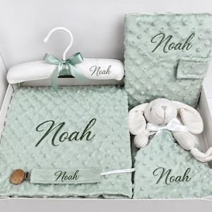 Op de afbeelding: Een saliegroene babycadeauset met een deken met noppenstructuur, een knuffeldoekje in de vorm van een konijn, een documentenmap en een witte gevoerde kledinghanger, allemaal geborduurd met de naam Noah, plus een saliegroene speenkoord met de naam Noah.