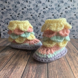 Könnte beinhalten: Ein Paar gehäkelte Babyschuhe mit einem mehrschichtigen Rüschen-Design in Pastellgelb, Rosa und Grün.