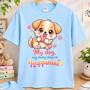 Könnte beinhalten: Ein hellblaues Kurzarm-T-Shirt mit einem Cartoon-Welpen, der ein rosa Herz hält, umgeben von kleinen Herzen und Sternen. Der Text My dog, my daily dose of happiness ist in rosa und gelben Farbverlauf-Buchstaben auf der Vorderseite gedruckt.