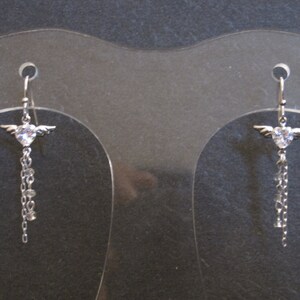 May include: A pair of silver dangle earrings featuring a clear crystal heart with wings. The earrings have a chain with small clear crystals.