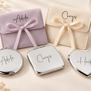 Pode incluir: Duas bolsas de veludo macio em lavanda e creme, cada uma amarrada com uma fita correspondente e personalizada com os nomes Adele e Carys. Abaixo, três espelhos compactos de prata polida em formas redonda, quadrada e de coração, gravados com Adele, Carys e Heidi.