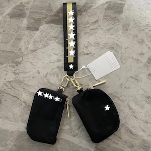 Op de afbeelding: Twee zwarte etuis met ritssluiting bevestigd aan een zwart-gouden polsband. De band heeft een verticale rij witte sterren. Eén etui heeft vier witte sterren en de tekst "lmn" erop, de andere heeft een enkele witte ster.
