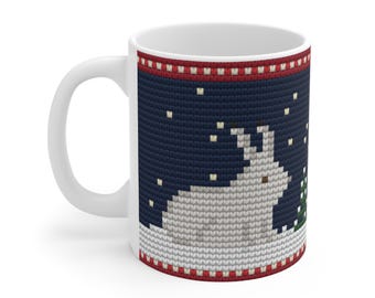 May include: A white ceramic mug featuring a printed design of a light gray rabbit sitting in snow next to a small green pine tree against a dark blue background, framed by red and white patterned borders resembling a knitted sweater.