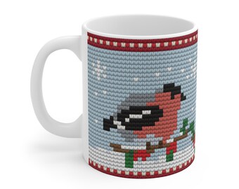 May include: A white ceramic mug featuring a printed pattern that mimics a knit texture. The design displays a bullfinch bird perched on a branch against a light blue background with white snowflakes, framed by red and white decorative borders.