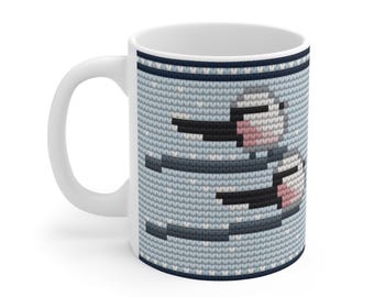 May include: A white ceramic mug featuring a repeating knit-texture pattern in light blue, dark blue, and white. The design displays a stylized bird with a long black beak, pink cheeks, and dark wings, set against a background of small white dots.