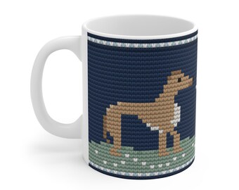 May include: A white ceramic mug featuring a dark blue background with a knitted-style pattern. The design displays a tan dog standing on a light green grassy area, framed by a decorative border of light blue and white stitches at the top and bottom.