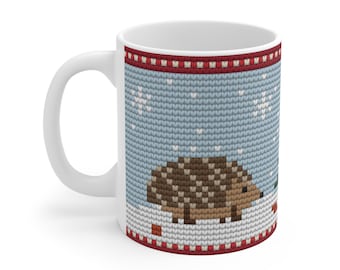 May include: A white ceramic mug featuring a printed pattern that mimics a knitted texture. The design displays a brown hedgehog standing on white snow against a light blue background with white snowflakes and red borders.