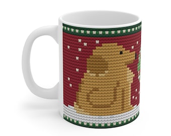 May include: A white ceramic mug featuring a red background with a knitted-style pattern. The design displays a tan dog sitting in the snow, a green wreath, and small white dots representing falling snow, framed by green and white borders.