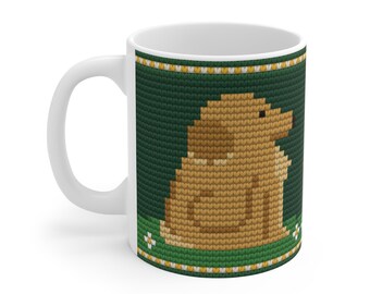 May include: A white ceramic mug featuring a dark green background with a knitted texture pattern. The design displays a light brown dog sitting in profile, surrounded by small white flowers and a yellow and white border at the top and bottom.