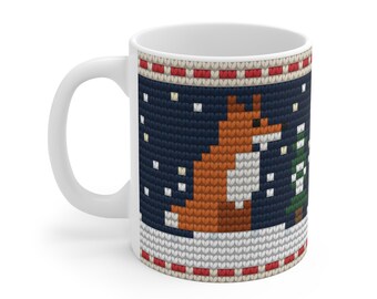 May include: A white ceramic mug featuring a printed pattern that mimics a knitted texture. The design displays a fox sitting in a snowy landscape with a small green tree against a dark blue background, framed by red and white borders.