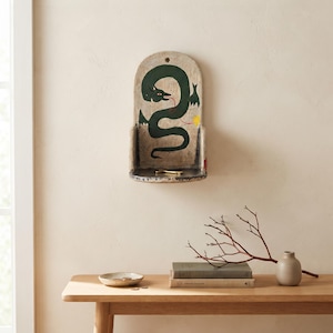Puede incluir: Un estante de madera montado en la pared con la parte superior redondeada, que presenta un dragón verde oscuro pintado a mano sobre un fondo blanco roto desgastado. El estante tiene una pequeña superficie plana y un orificio para colgar.