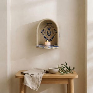 May include: An arched wall-mounted shelf featuring a beige finish with a blue floral illustration on the back panel. The shelf surface displays a blue and white checkered pattern. A small white tea light candle sits centered on the shelf surface.