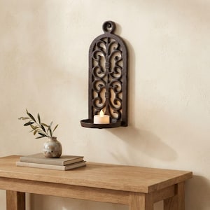 May include: A dark brown, arched wall-mounted candle holder featuring an intricate, openwork scroll design. The piece includes a circular base at the bottom that supports a single, small, white, battery-operated tea light candle with a warm glow.