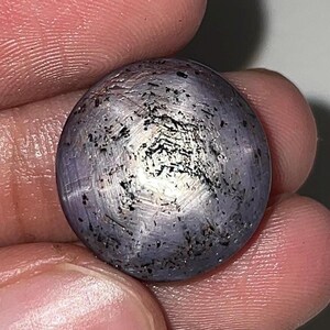 Puede incluir: Una piedra preciosa de zafiro estrella tallada en cabujón redondo con un tono grisáceo-púrpura translúcido. La superficie muestra un efecto de estrella de seis rayos centrado en la cúpula, junto con numerosas inclusiones oscuras en forma de aguja.