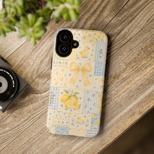 May include: A smartphone case featuring a patchwork design with soft yellow and blue hues. The pattern includes illustrations of small yellow ducklings, a large central yellow bow, lemons with green leaves, and various floral motifs.