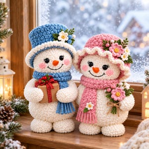 May include: Two crocheted snowmen figures. The left snowman wears a blue hat and scarf, holding a small gift box. The right snowman wears a pink hat and scarf, holding a bouquet of pink and white flowers. Both have orange noses and rosy cheeks.